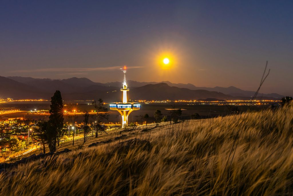 Bright tower near grass and street lights under sky with glowing sun illuminating mountains in twilight